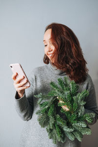 Adult woman with diy fir christmas wreath in hand using mobile phone on the grey background