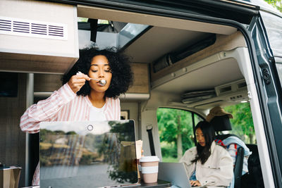 Young black woman preparing coffee while watching video on laptop inside camper vehicle with asian girlfriend during summer journey in nature