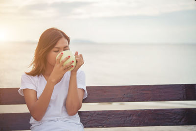 Woman drinking coffee while sitting on wooden swing at beach against sky during sunset