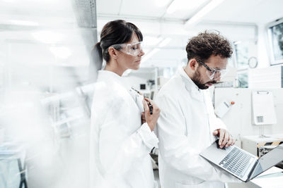 Female scientist standing by male colleague using laptop at laboratory