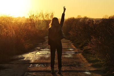Rear view of woman gesturing peace sign while standing on road against sky during sunset