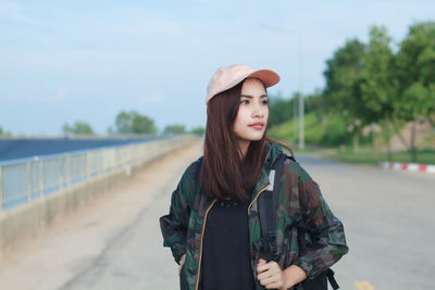Young woman looking away while standing on road against clear sky