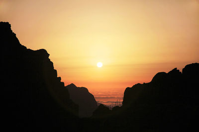 Silhouette cliff by sea against orange sky