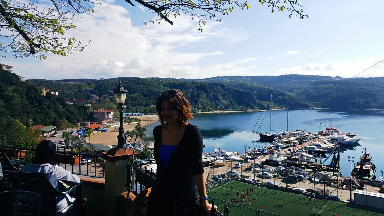 lifestyles, water, leisure activity, sky, casual clothing, standing, person, young adult, three quarter length, lake, mountain, tree, young women, river, sitting, railing, waist up, cloud - sky