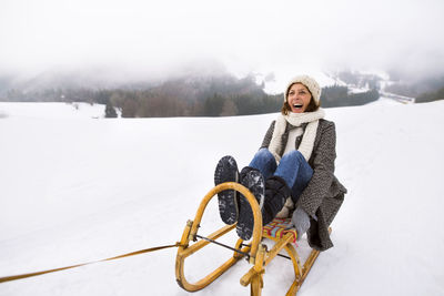 Laughing senior woman sitting on sledge in snow-covered landscape