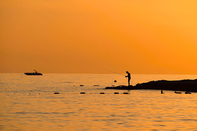 Silhouette man fishing in sea against orange sky