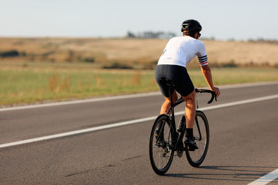 Back view of professional cyclist in sport clothing and helmet biking on asphalt road. 