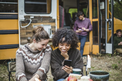 Smiling female friends sharing smart phone while sitting against caravan during camping