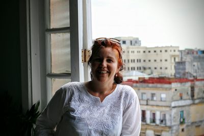 Portrait of happy woman leaning on window in city against clear sky