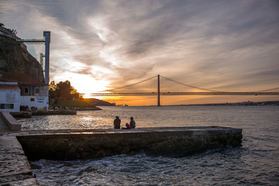 People on suspension bridge over sea against sky during sunset