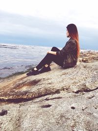 Side view of young woman sitting on rock at beach against sky