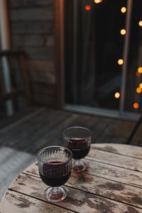 Two glasses of red wine standing on the wooden table with blurred trees and lights 