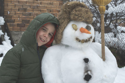 Portrait of smiling boy by snowman in yard during winter