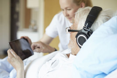 Senior man wearing headphones while using digital tablet with female nurse on hospital bed