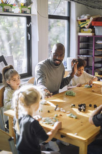 Male teacher playing with kids while sitting at bench during art class at preschool