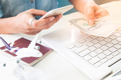 Midsection of woman holding currencies while using mobile phone with laptop on table