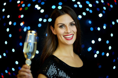 Portrait of beautiful young woman holding champagne flute against illuminated lights