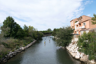 People by river amidst trees and buildings against sky