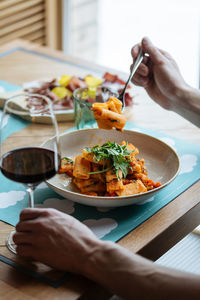 Cropped unrecognizable person eating tasty vegan pasta decorated with fresh rocket salad leaves and sauce and drinking red wine in a glass at a restaurant table