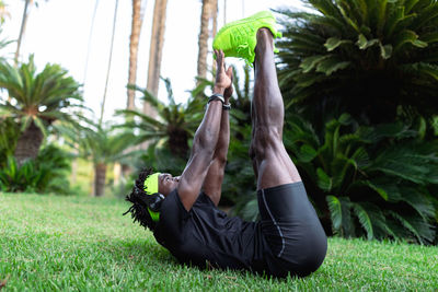 Young african american sportsman in stylish activewear sitting on green grass and doing stretching exercises after running in park