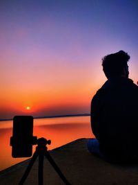 Silhouette photographing camera on sea against sky during sunset