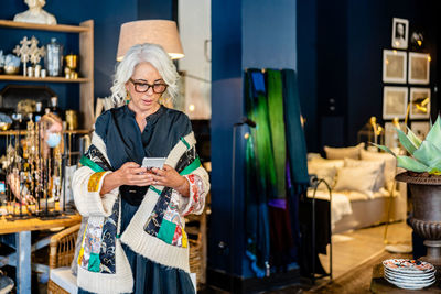 Elegant middle aged female in stylish outfit and glasses messaging on mobile phone while spending time at home with vintage interior