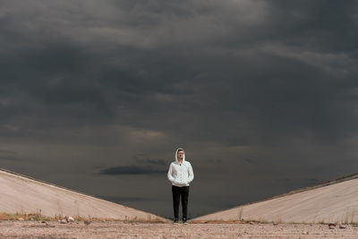 Portrait of young man wearing hooded shirt standing on field against cloudy sky