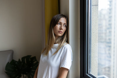 Portrait of young woman looking through window at home