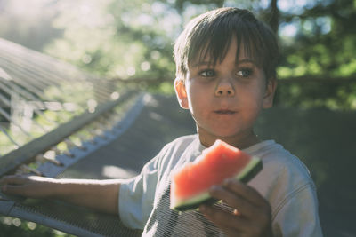 Boy eating watermelon against hammock during sunny day