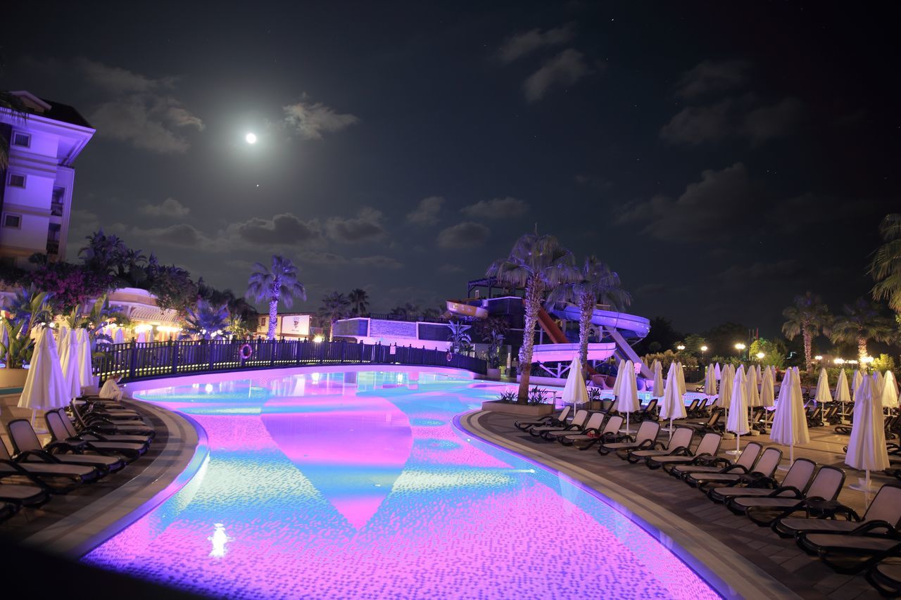 illuminated, night, water, pool, swimming pool, sky, built structure, architecture, nature, building exterior, incidental people, lighting equipment, dusk, table, seat, travel destinations, cloud - sky, chair, outdoors, luxury, purple