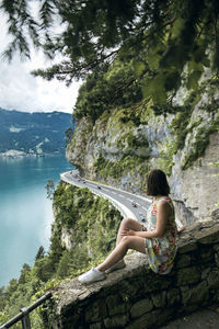 Side view of woman sitting on retaining wall over road by lake