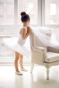 Little ballerina girl 2 in the studio in white tutu dress clothes, chair and looking out the window