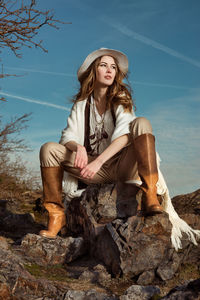 Fashionable young woman sitting on rock against sky