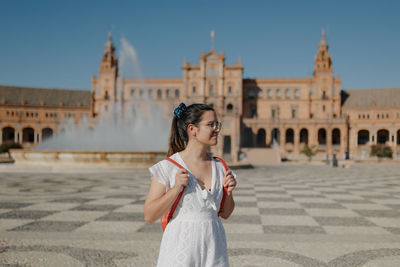 Young tourist woman wearing a white dress is looking away and smiling while standing in seville.