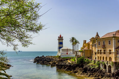 Lighthouse and buildings by sea against clear sky