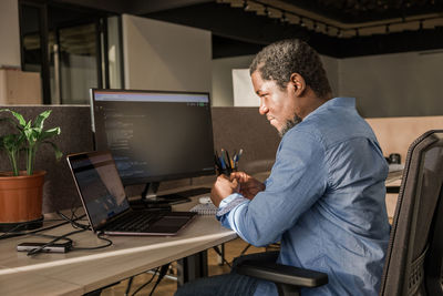 Black man writing lines of code on desktop pc with two monitors and a laptop aside in stylish office