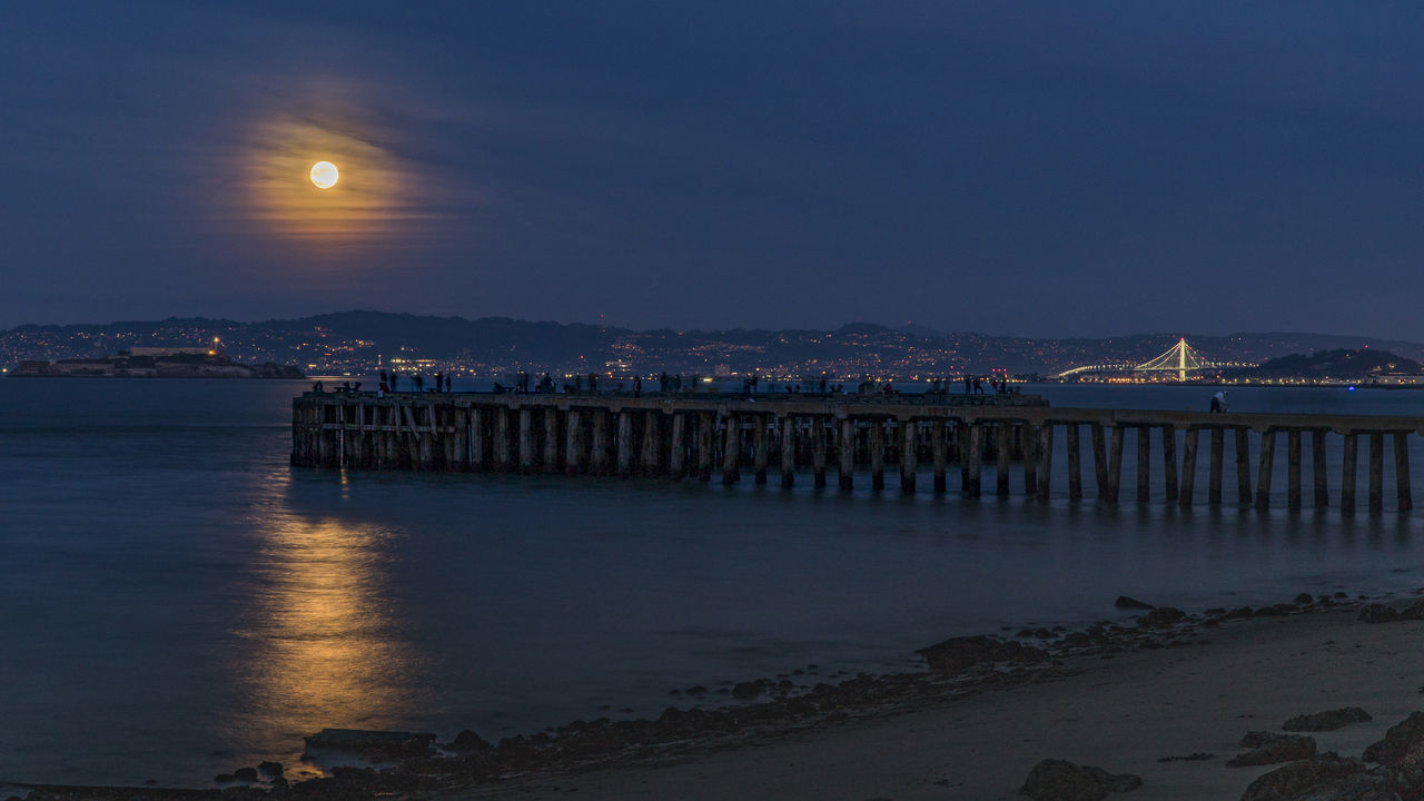 ILLUMINATED BRIDGE OVER SEA AGAINST SKY AT SUNSET