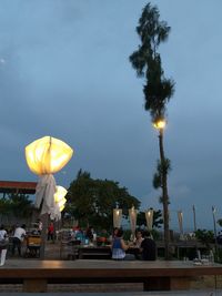 People sitting by illuminated street light against sky at dusk