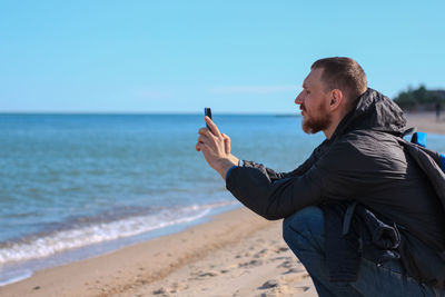 Bearded man in black raincoat on blue sea landscape background with smartphone in hands takes selfie