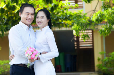 Portrait of smiling young couple with flower bouquet standing against building