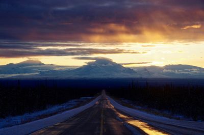 Empty road leading towards mountains against sky at sunset