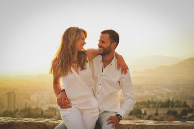 Smiling young couple sitting against townscape on retaining wall