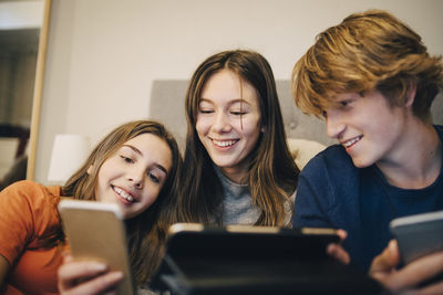 Close-up of happy friends using technology while sitting on bed at home