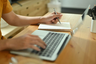 Cropped image of a hand taking notes at the wooden table surrounded by a laptop and pencil holder.