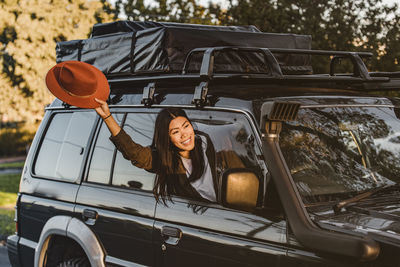 Positive asian female traveler peeping out of car window with outstretched arm and waving hand while enjoying vacation in australia