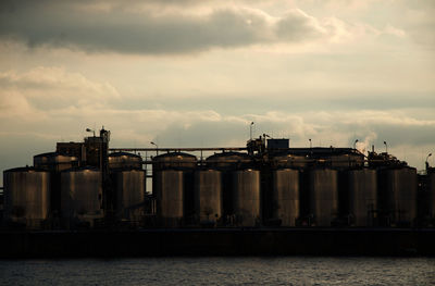 Industry by river against sky during sunset