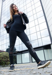 Low angle portrait of young woman standing on footpath against building