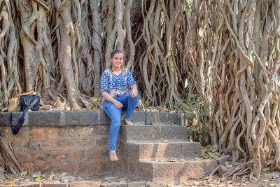 Full length portrait of smiling woman sitting against tree