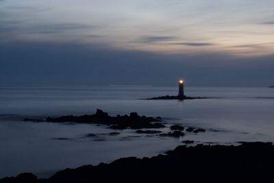 Silhouette lighthouse by building against sky at sunset