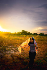 Young woman standing with sarong on field against sky during sunset